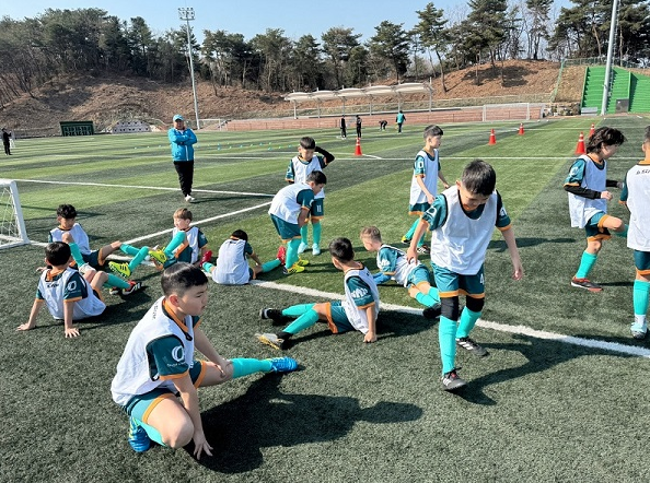 Young children in soccer jerseys and shorts, some stretching or warming up on a soccer field during practice or training session, with coaches and orange cones in the background.