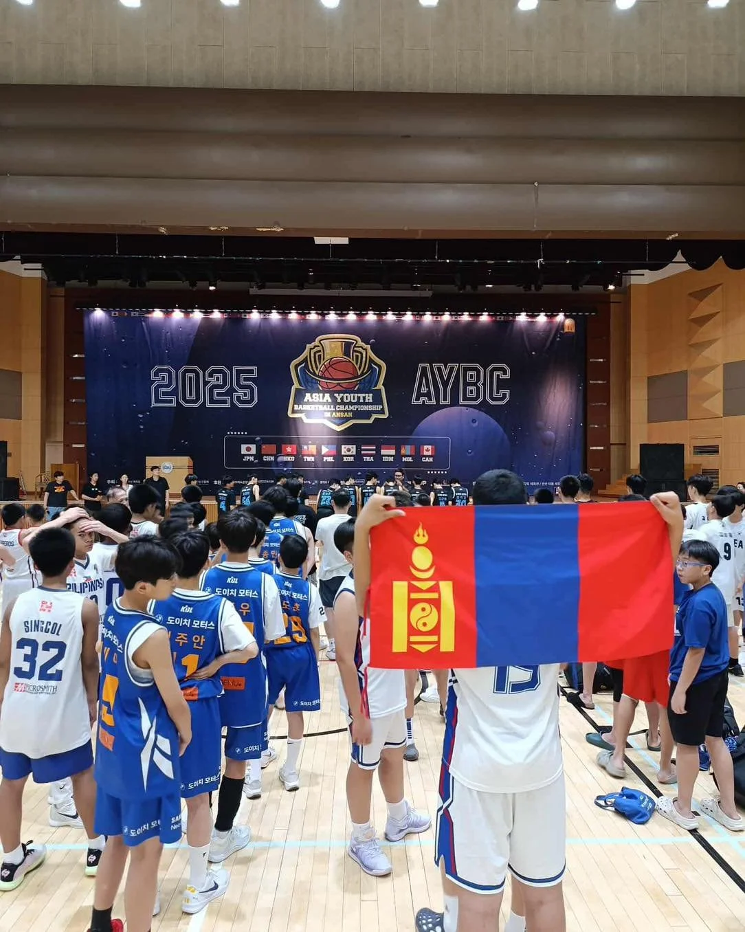 Young basketball players in uniforms gathered in a gymnasium watching a ceremony on stage for the 2025 Asia Youth Basketball Championship, with a person holding a flag in the foreground.