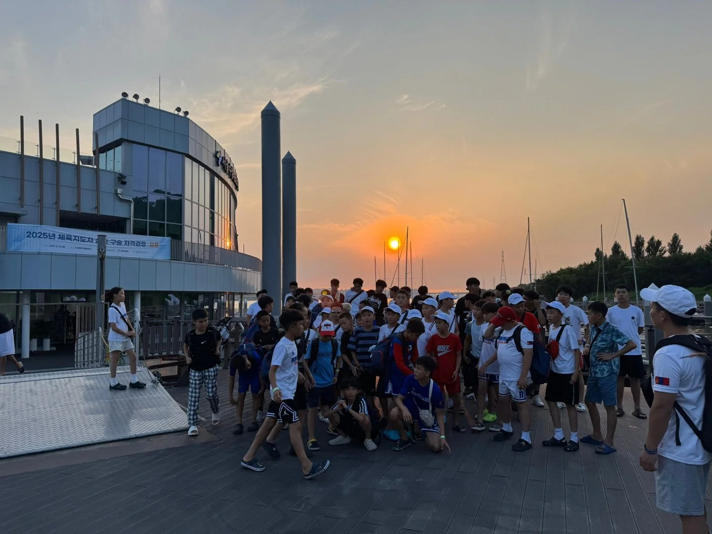 Group of children and teenagers gathered on a dock during sunset, near boats and a modern building.