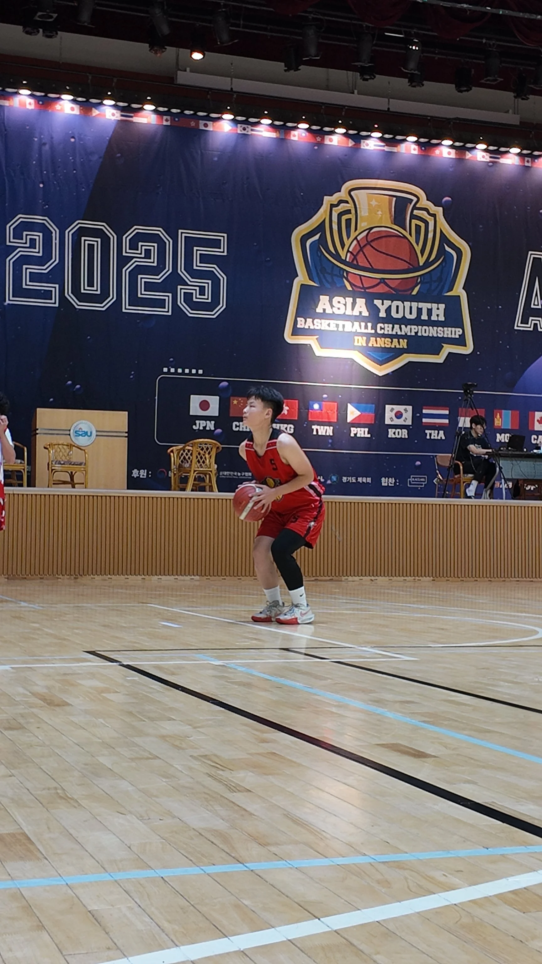 A young basketball player in a red uniform preparing to shoot on an indoor court during the Asia Youth Basketball Championship in Ansan, with a large banner displaying flags and the event logo in the background.