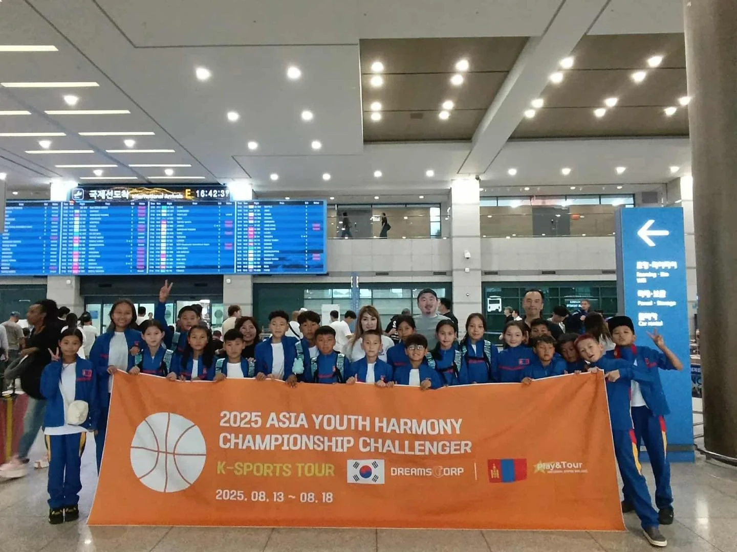 Group of children and adults holding an orange banner at an airport, with flight information display in the background. The banner reads '2025 Asia Youth Harmony Championship Challenger, K-Sports Tour, August 13-18, 2025', and features logos of South Korea, Dreams Corp, and Play&Tour.