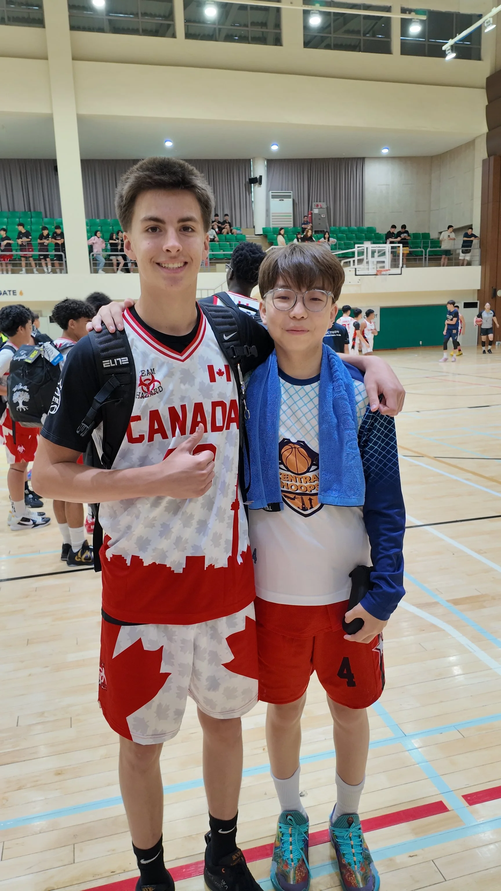 Two boys in basketball uniforms standing on an indoor basketball court with their arms around each other, smiling at the camera. The taller boy is wearing a Canadian basketball jersey and shorts, giving a thumbs-up. The shorter boy is wearing a baske