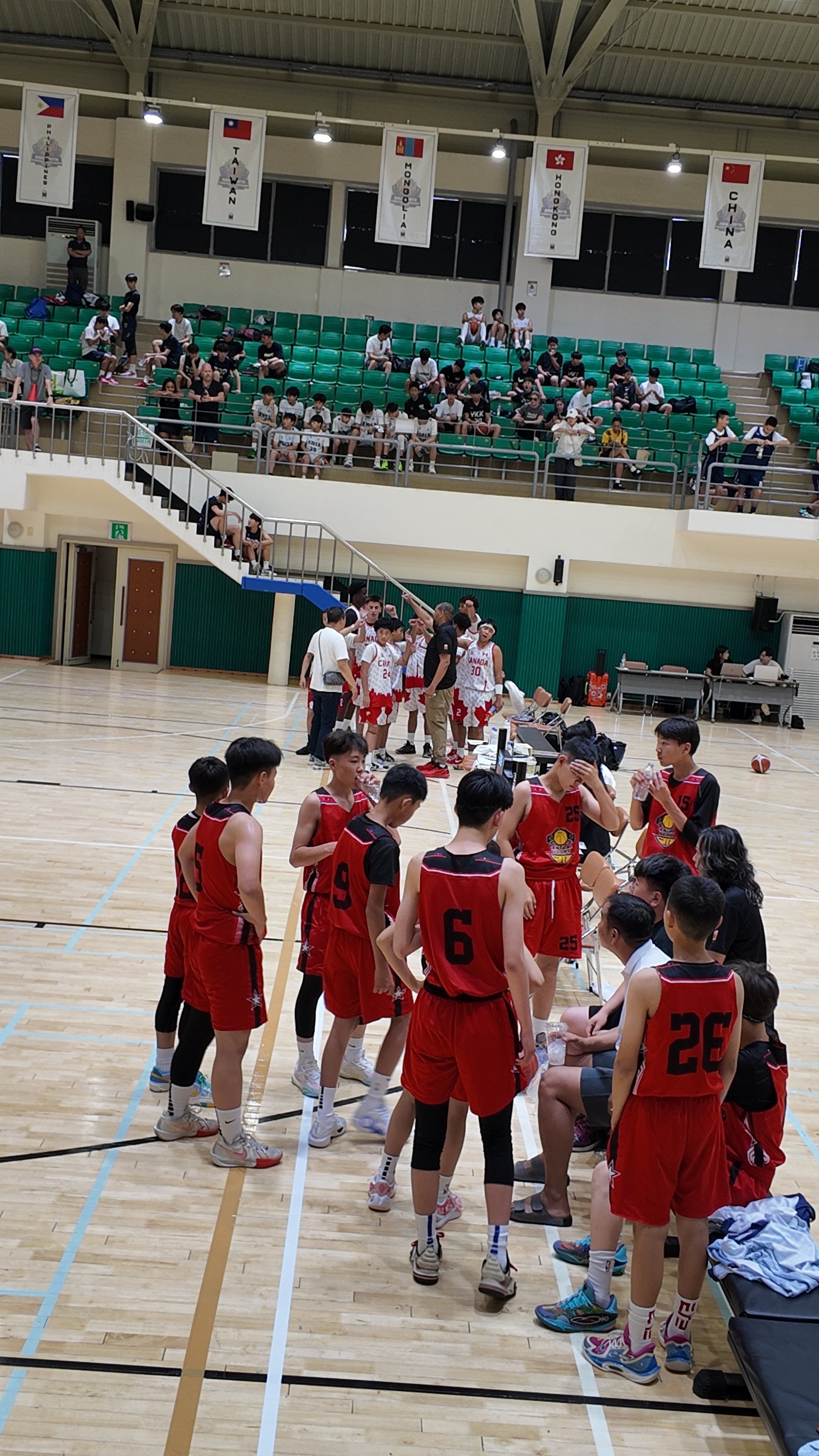 A basketball team in red uniforms is gathered around a coach during a timeout on the court, with players standing and sitting, while the spectators watch from the bleachers in the background.