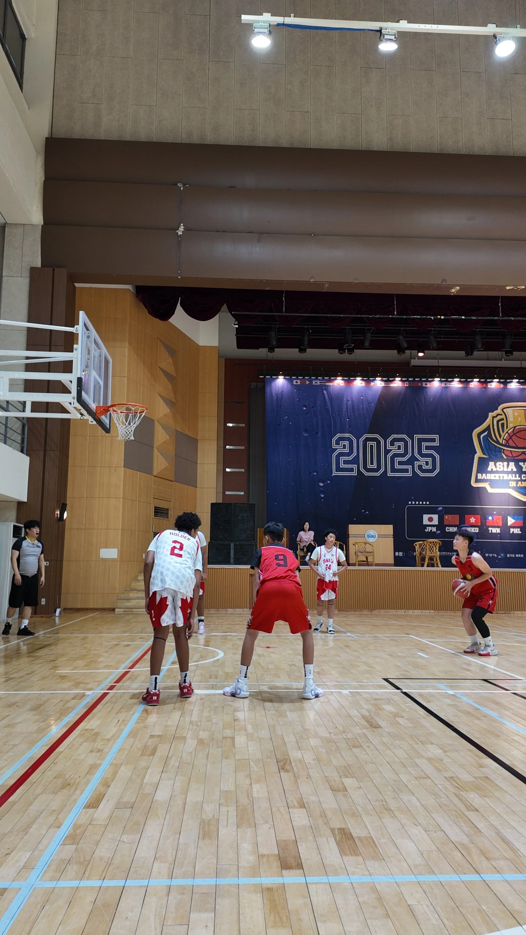 Youth basketball game with players in red and white jerseys practicing free throws on an indoor court, with a stage and large banner displaying '2025' in the background.