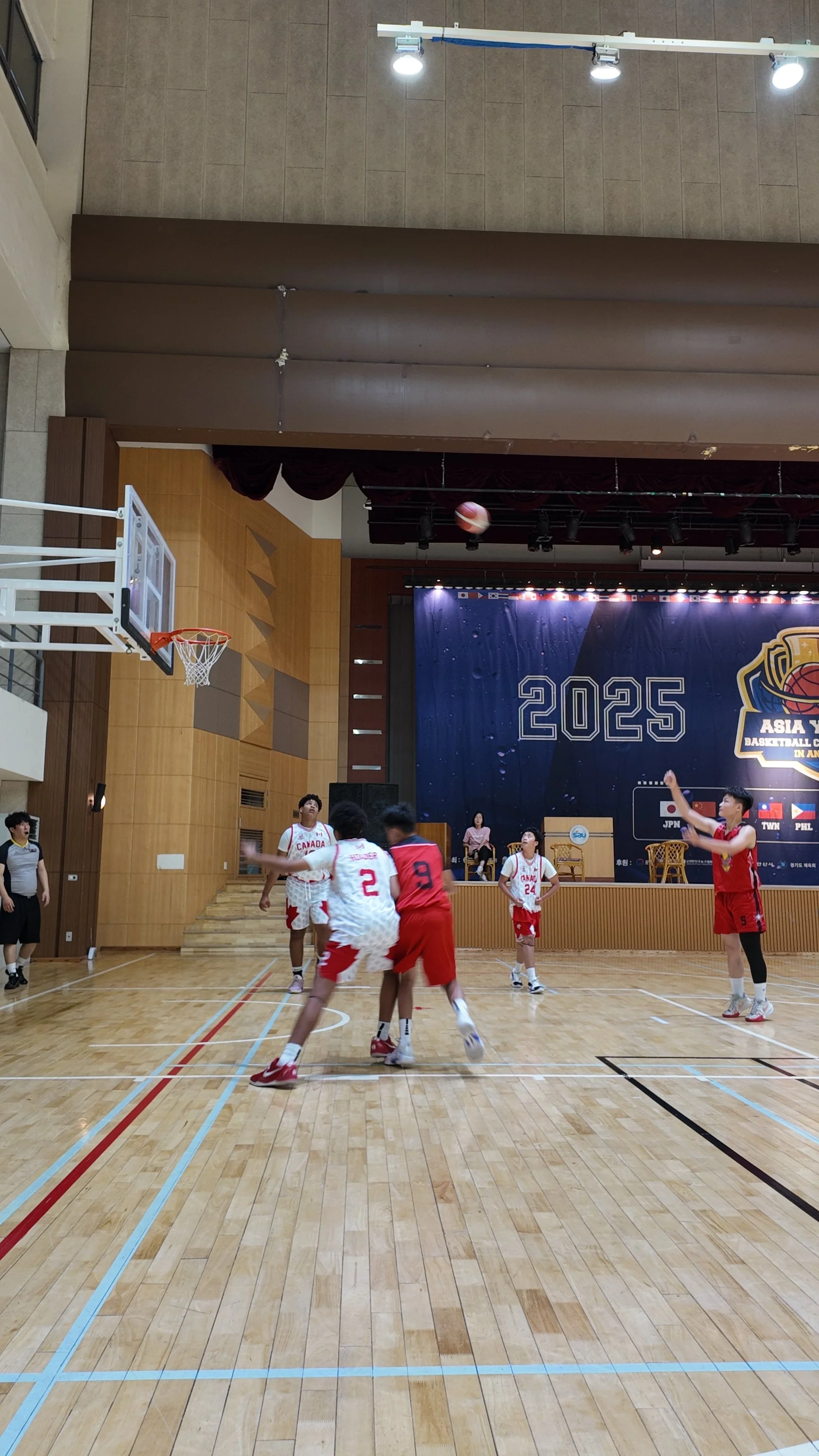 Young basketball players practicing on an indoor court during a game at the 2025 Asia Youth Basketball Championship, with players in white and red jerseys, a referee, and a referee's assistant.
