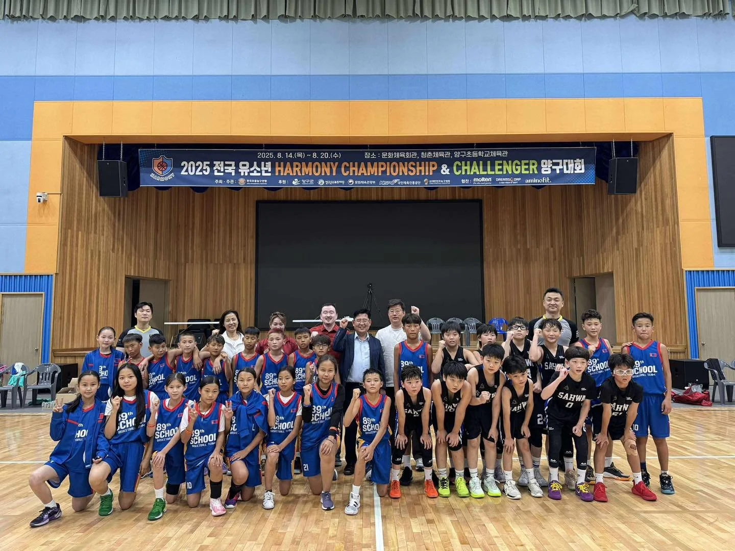 Group of young basketball players in blue and black uniforms posing for a photo on a basketball court, with adults standing behind them and a banner overhead that reads '2025 전국 유소년 Harmony Championship & Challenger'.