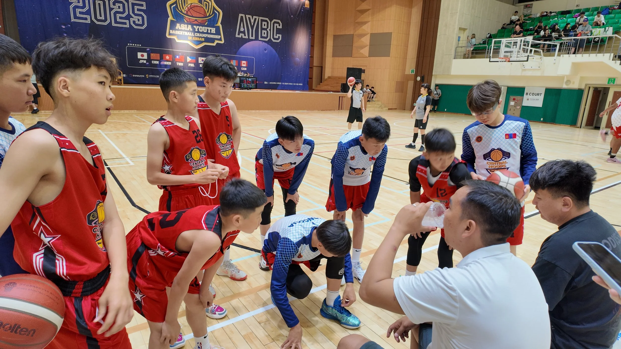Young male basketball players in red and blue uniforms huddle and listen to coaches during a timeout at an indoor basketball court, with some players in a crouched position and others standing, a basketball and water bottle are visible.