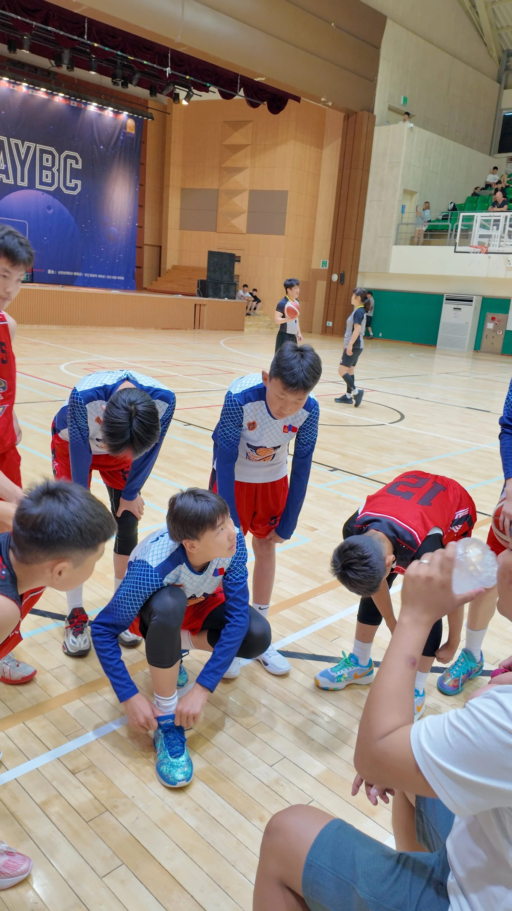 Children in basketball uniforms on an indoor basketball court, some kneeling, some standing, with a stage and large scoreboard in the background.