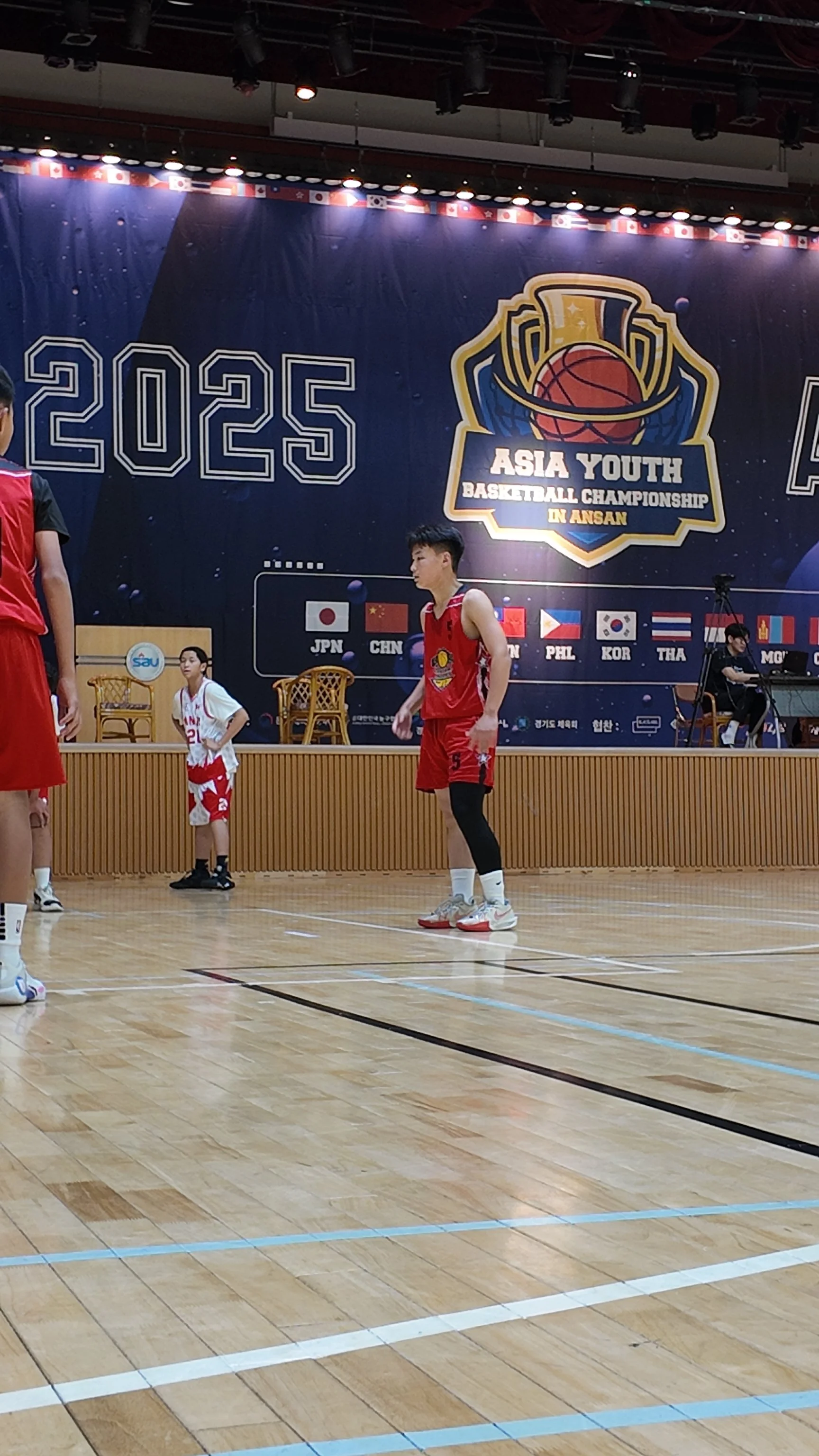 Young basketball players in red uniforms warming up on a wooden court during the Asia Youth Basketball Championship in Ansan, with a large backdrop displaying the event name and participating country flags.