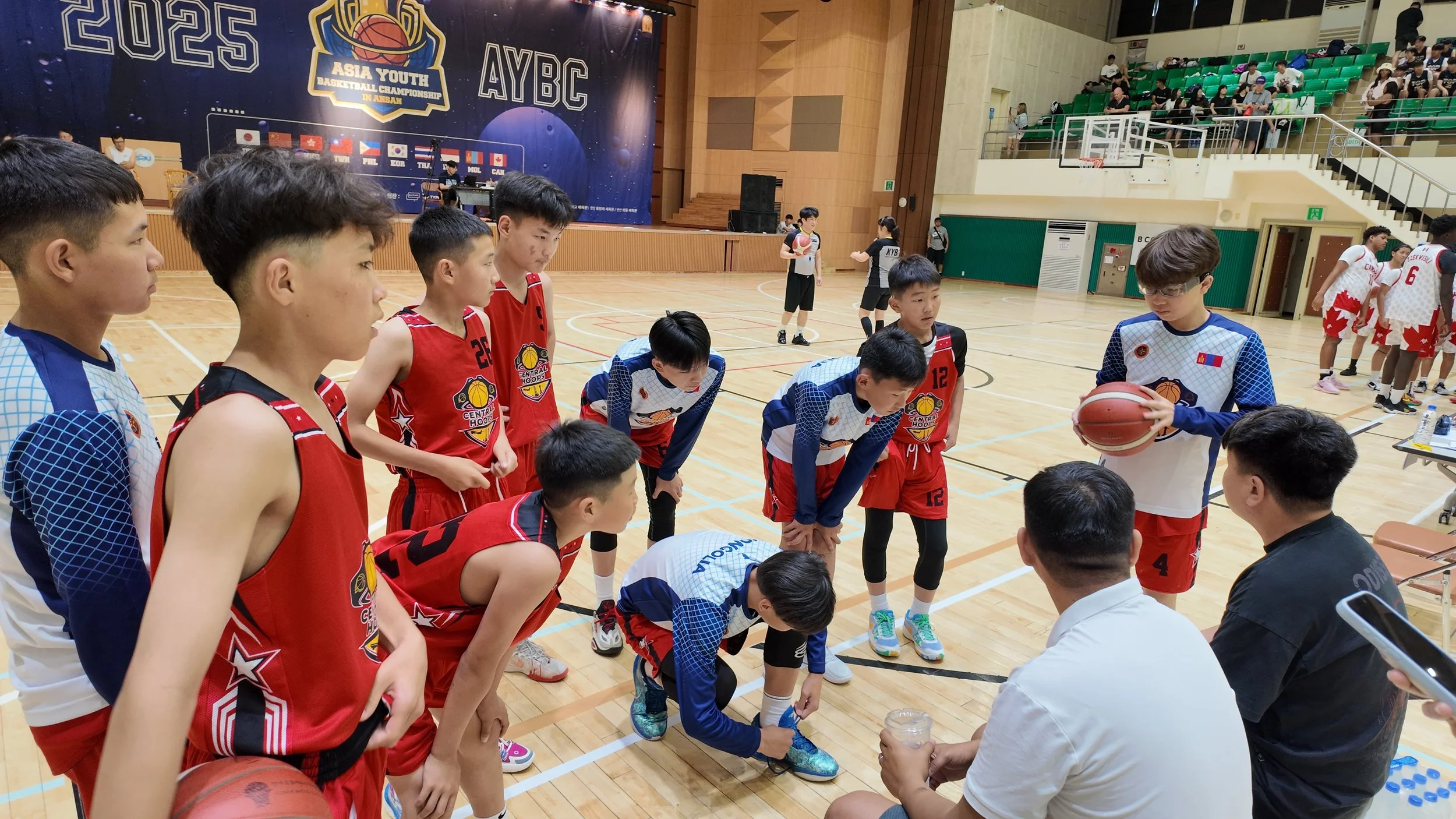 Young basketball players in red and blue uniforms listening to coaches during a timeout at the Asia Youth Basketball Championship in Ansan, with spectators in the stands and other teams warming up on the court.