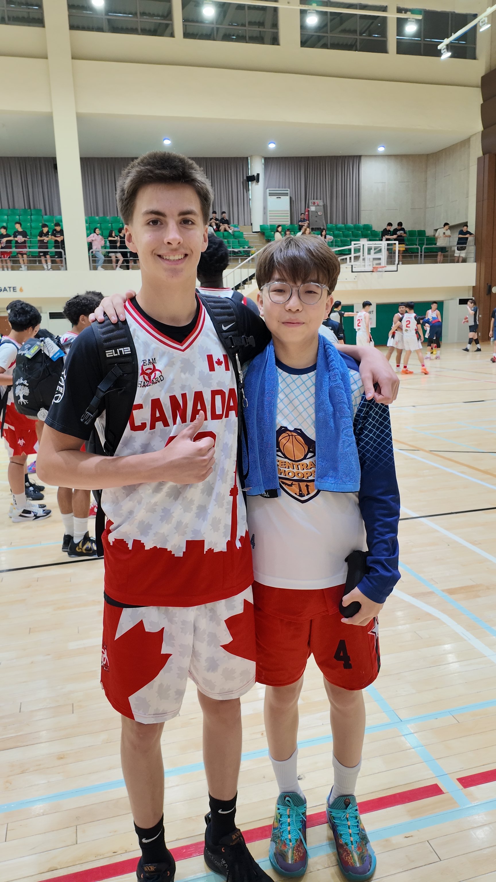 Two boys standing on a basketball court, smiling at the camera, with other players and spectators in the background.