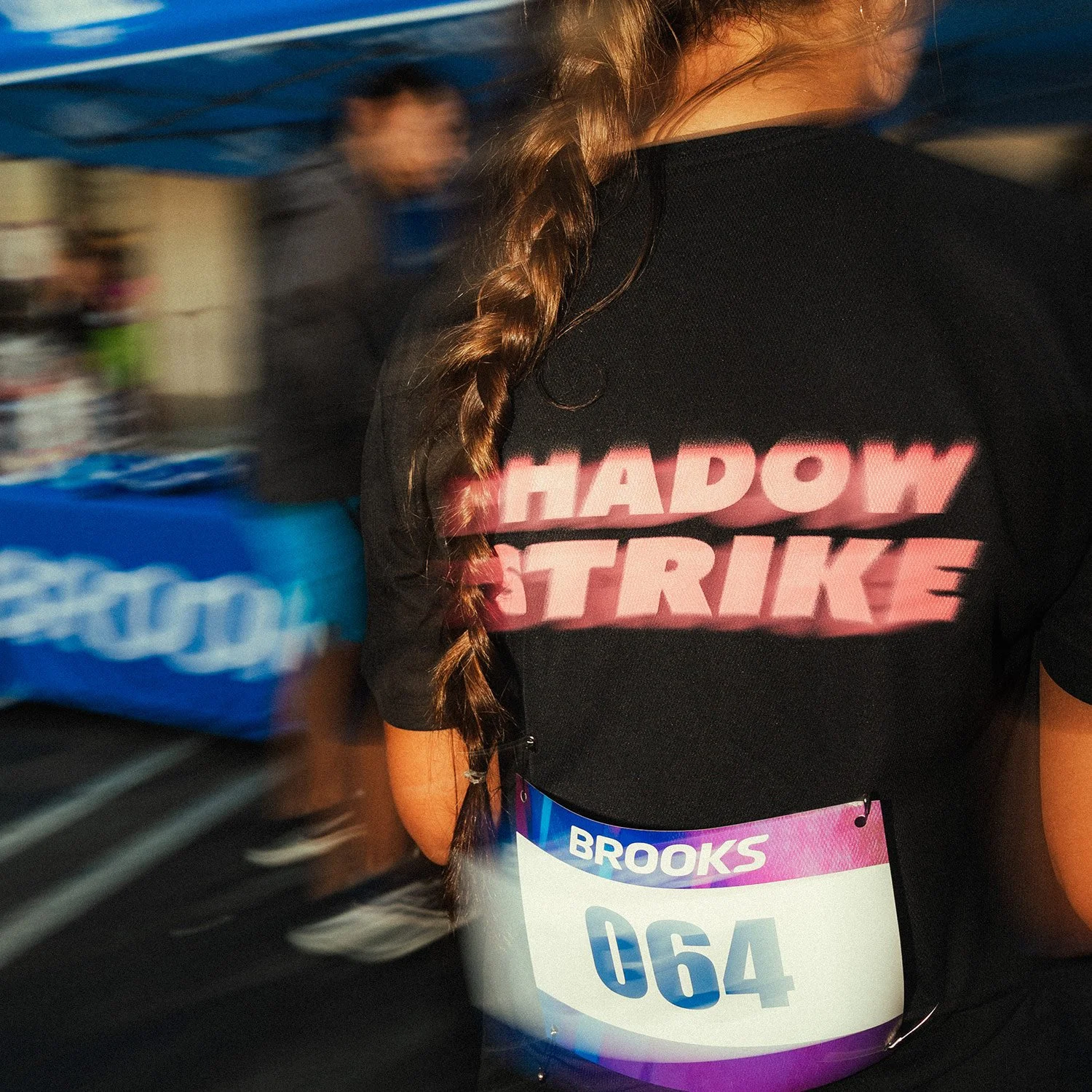 Back view of a Shadow Strike runner wearing an event shirt in Brea, CA, with a Brooks canopy in the distance offering race-day footwear to participants.