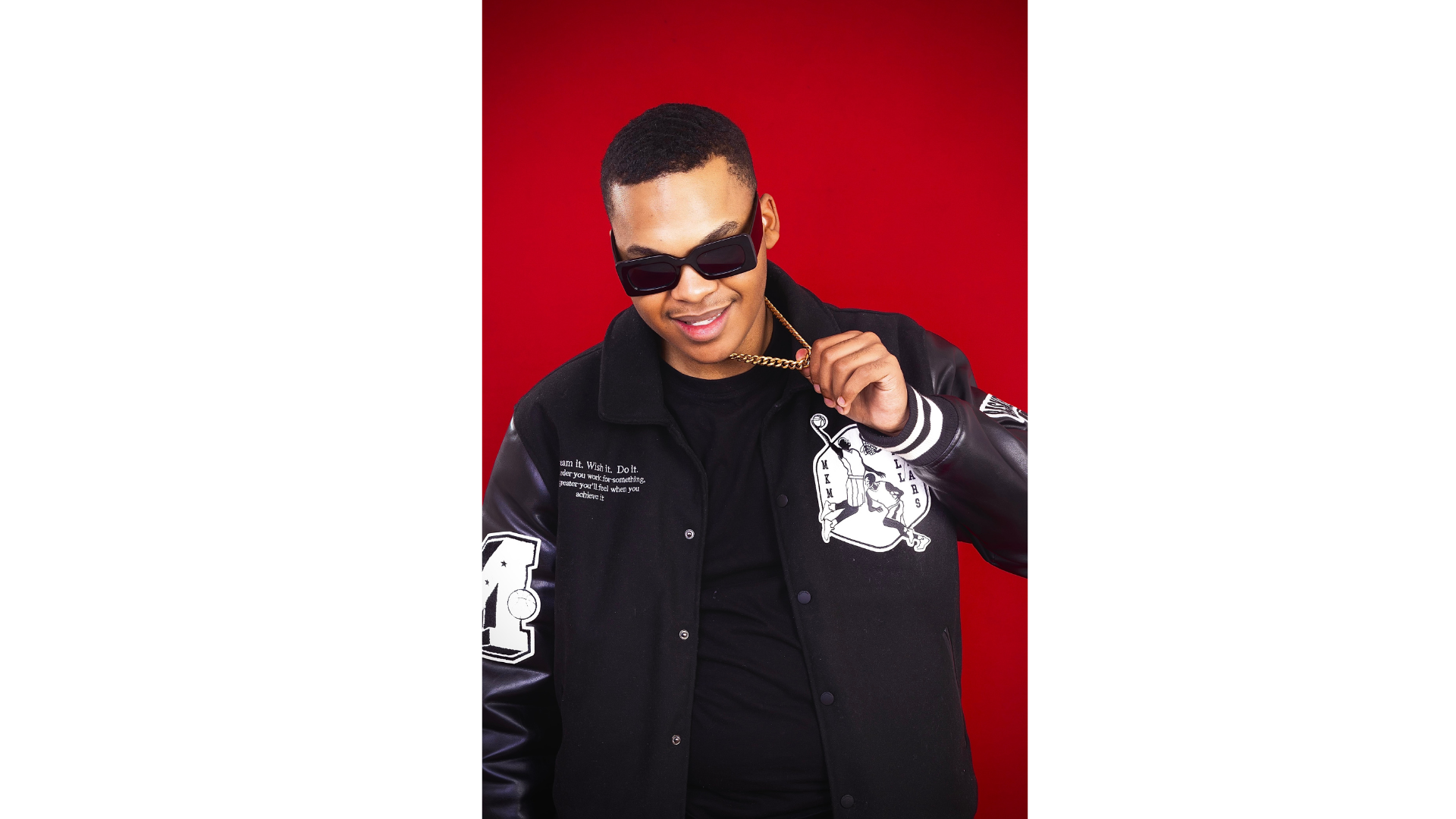Amari MustBeAmari Producer wearing sunglasses, black jacket, holding a gold chain, standing against a red background.