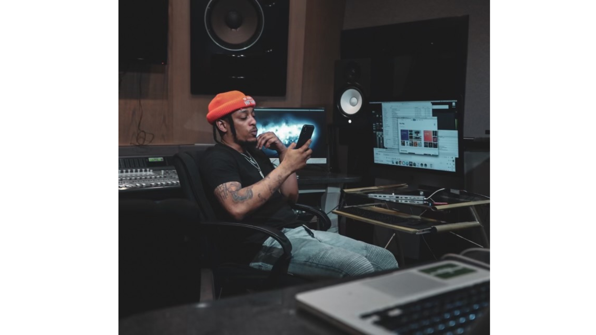 G-Elz in studio with orange beanie looking at phone, surrounded by audio equipment and computer screens.