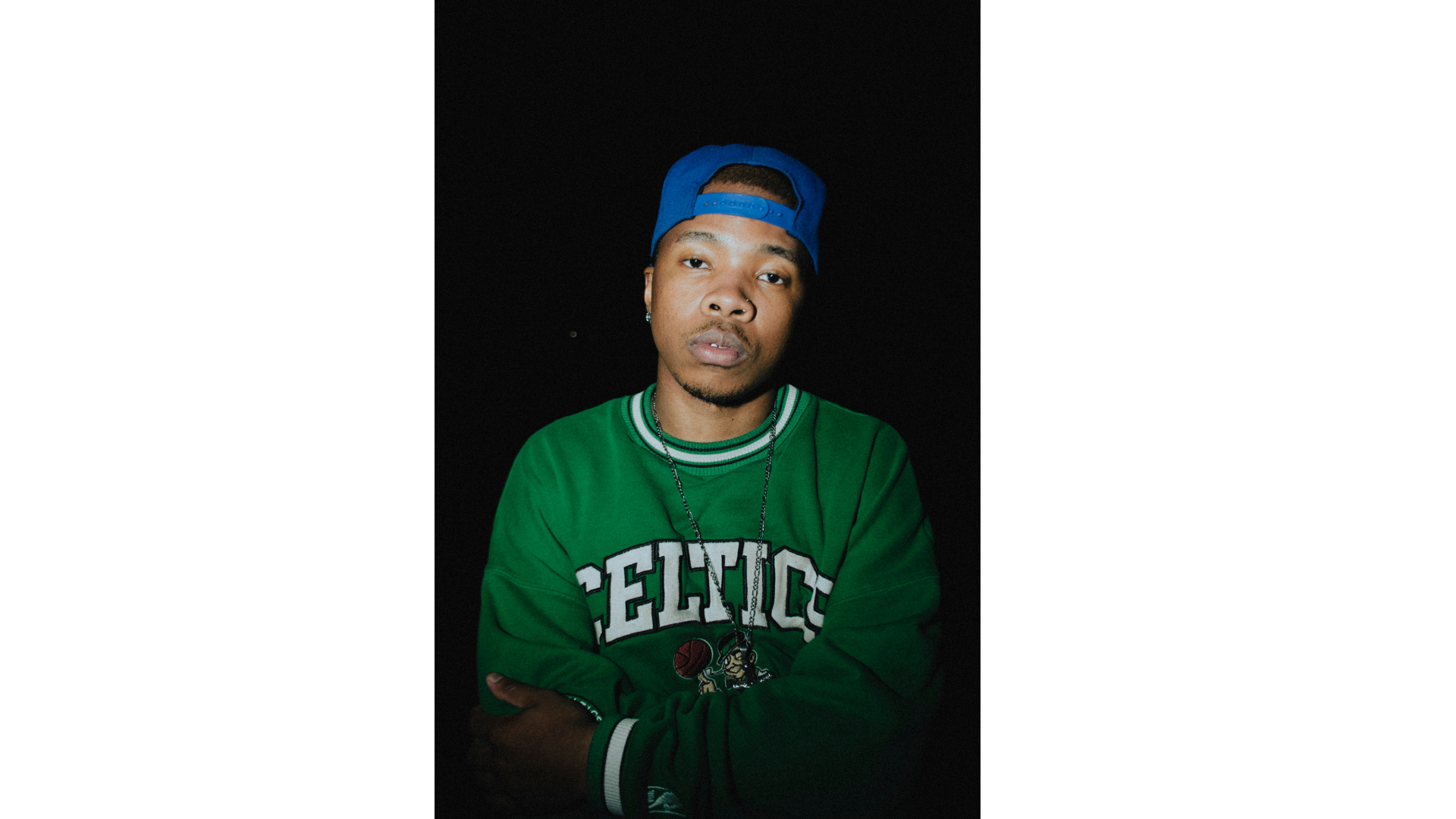 Lewi Ercoll rapper, singer Individual wearing a green Celtics sweatshirt and a blue cap against a dark background.