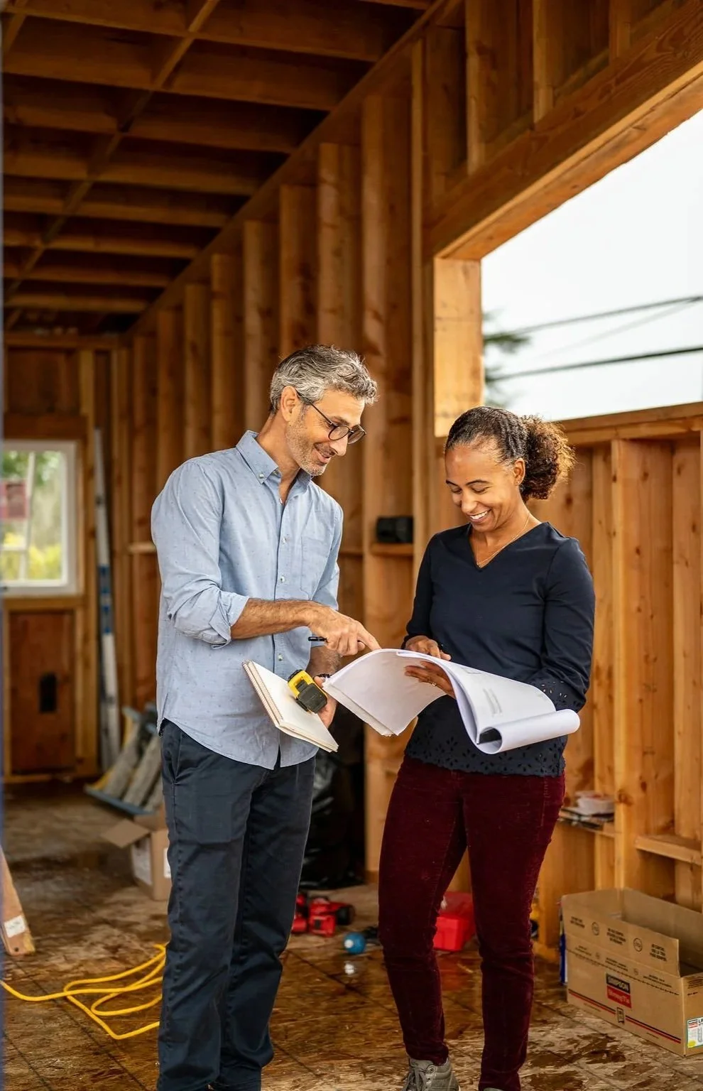 Two people, a man and a woman, discussing construction plans inside a wooden framing structure of a building under construction.