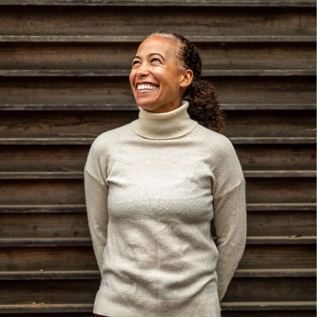 A woman with dark, curly hair tied back, wearing a light beige turtleneck sweater, standing outdoors in front of a wooden wall, smiling and looking upwards.