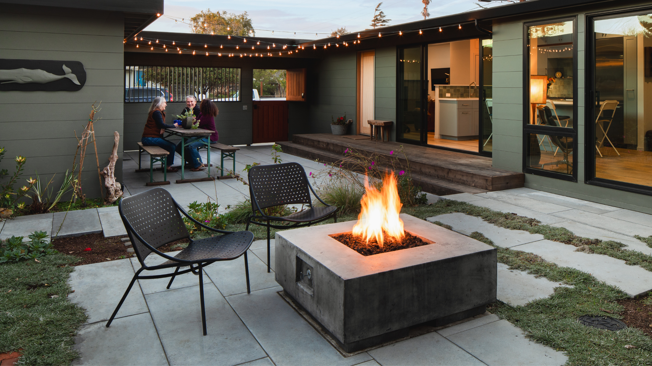 A backyard patio with a fire pit, two black chairs, and a group of three women sitting at a table. The house has large glass sliding doors and string lights overhead.