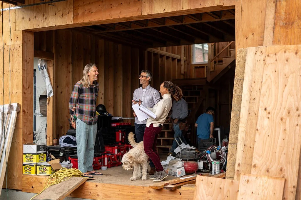 Group of people inside a house under construction, smiling and talking, with tools and materials around, including a dog.