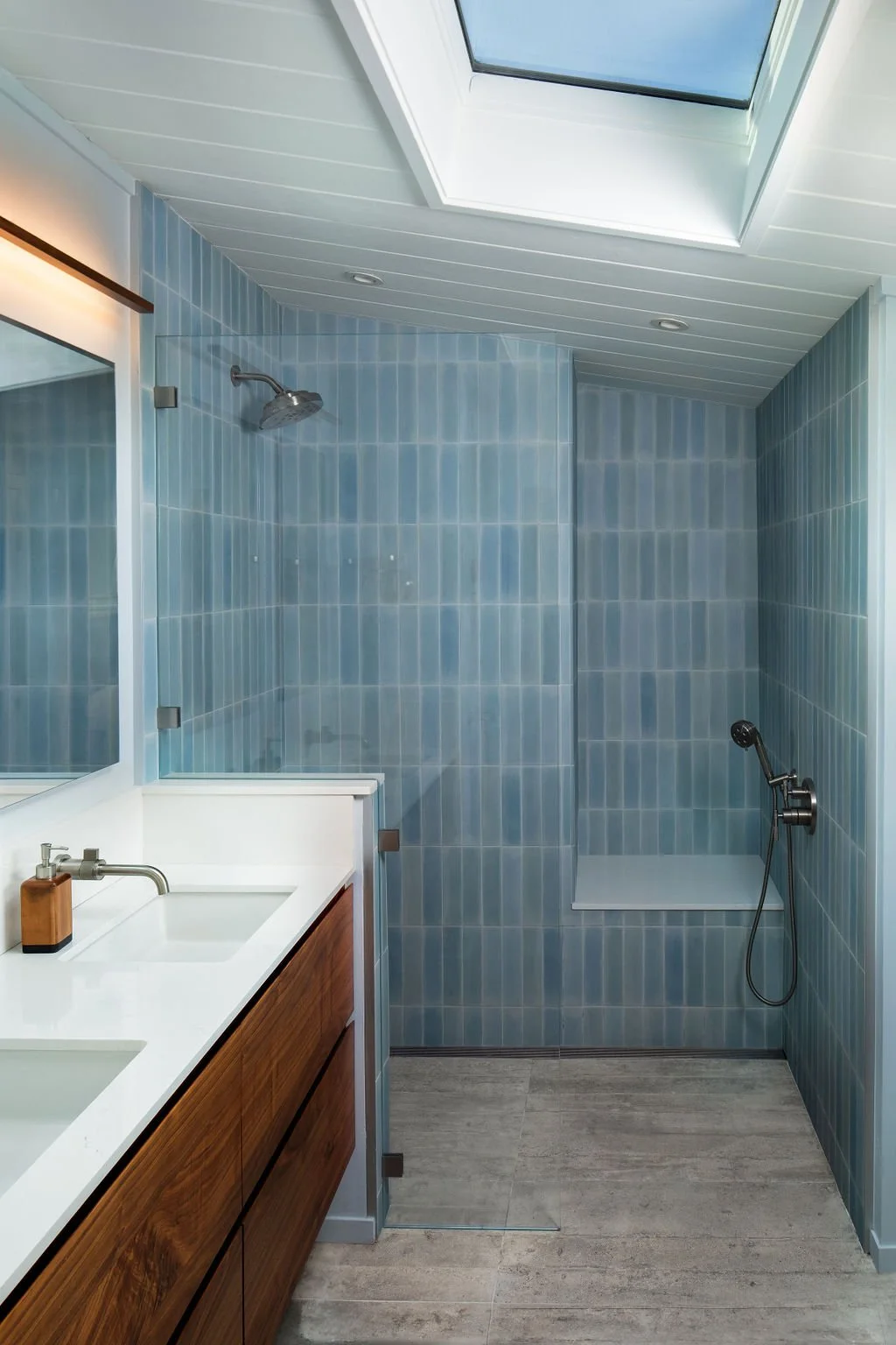 Modern bathroom with a glass-enclosed shower featuring blue tiles, a skylight, a white countertop with two sinks, and a wooden vanity.