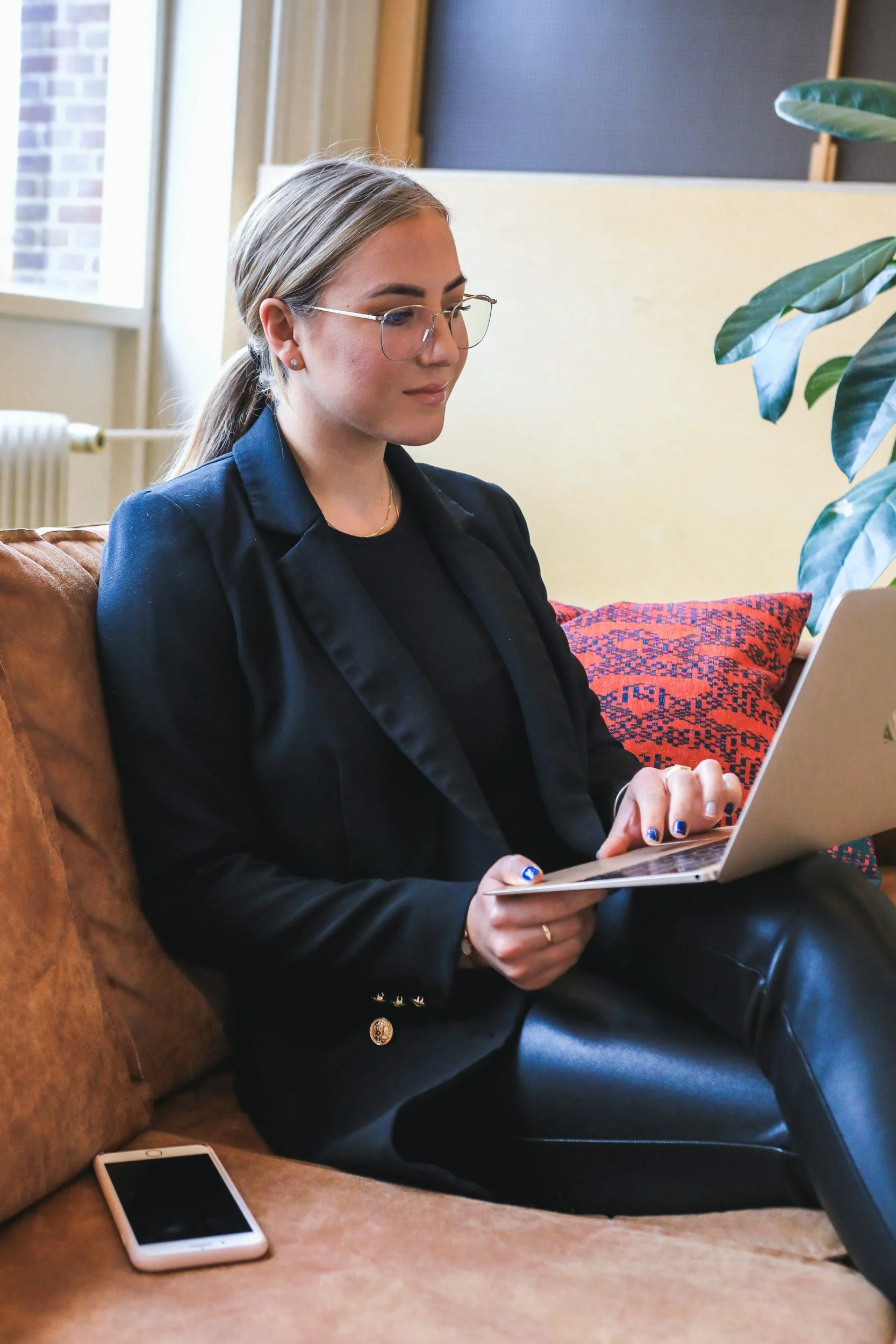 A woman with blonde hair tied back, wearing glasses, a black blazer, black shirt, and leather pants, is sitting on a brown couch searching for a bilingual job opportunity on a laptop in a well-lit room with large windows and a potted plant nearby.