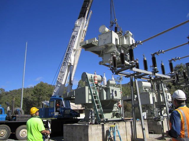 Construction workers using a crane to install electrical equipment at a power substation on a sunny day.