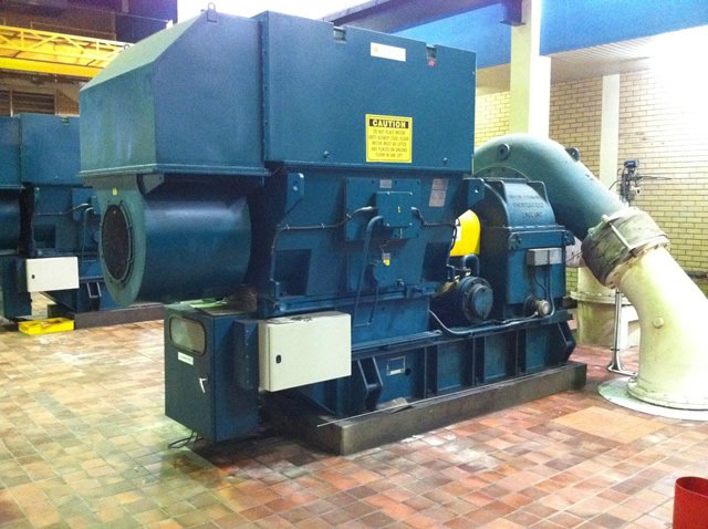 Industrial blue machinery in a tiled room with warning sign, connected to a large pipe.