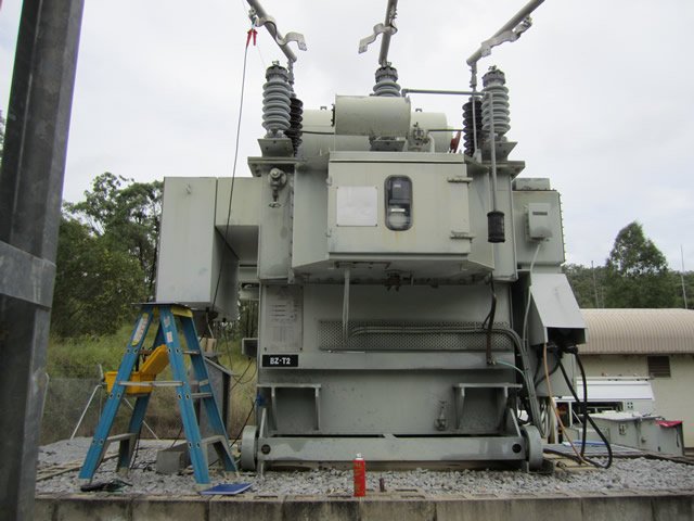 High-voltage electrical transformer outdoors on a concrete pad, with nearby equipment and a blue ladder.