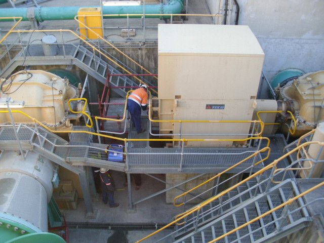 Industrial facility with machinery, pipes, and metal walkways, two workers in safety vests are inspecting equipment.