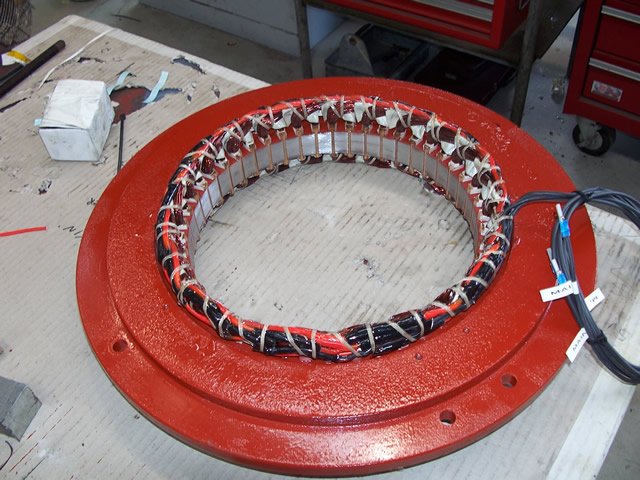 Red circular electric motor stator with visible copper windings and wiring, placed on a workbench.