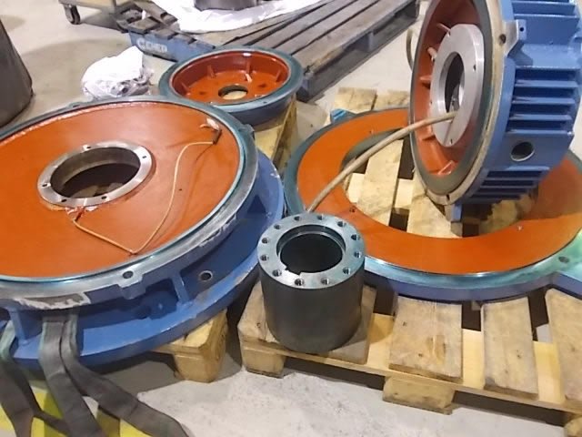Disassembled industrial machinery parts on a pallet, including large circular metal components with orange and blue surfaces, wires, and cylindrical pieces.