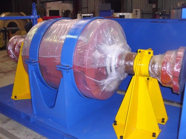 Large industrial cylinder wrapped in plastic, mounted on a blue and yellow support frame in a workshop.