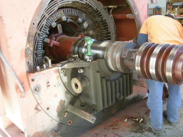 A technician performing maintenance on an industrial electric motor. The motor's rotor and stator are exposed, and various metal components are visible. The technician is wearing a safety vest and blue jeans.