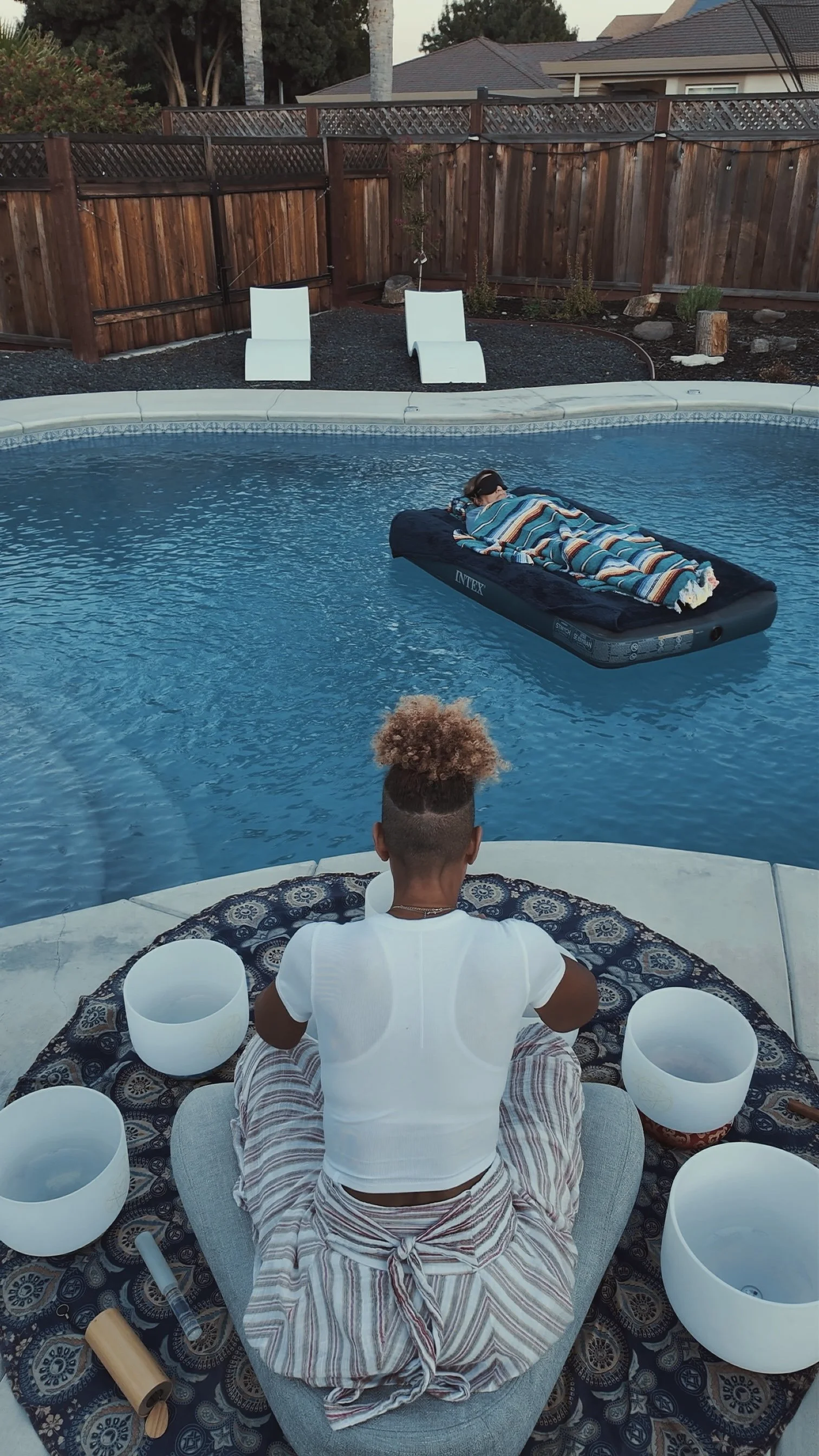 Person with curly hair colored at the sides, sitting cross-legged on a daybed by a pool, surrounded by white bowls, with a blanket, and other relaxing accessories, with a view of a pool and fencing in the backyard.