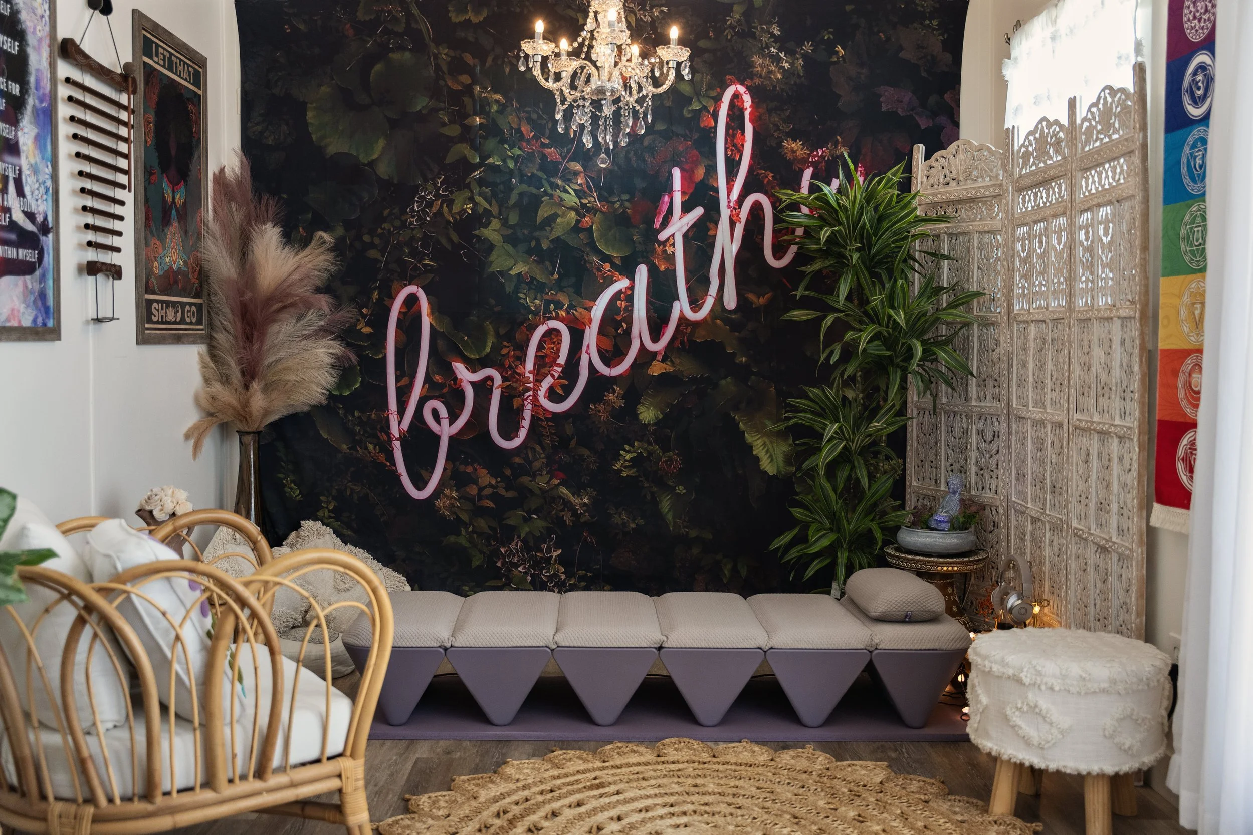 A bohemian styled living room corner featuring a dark leafy wall mural with a neon pink 'hello' sign, a small chandelier, a lush green plant, a textured cream sofa with cushions, a white ornate room divider, and a multicolored rainbow chakra hanging decoration.