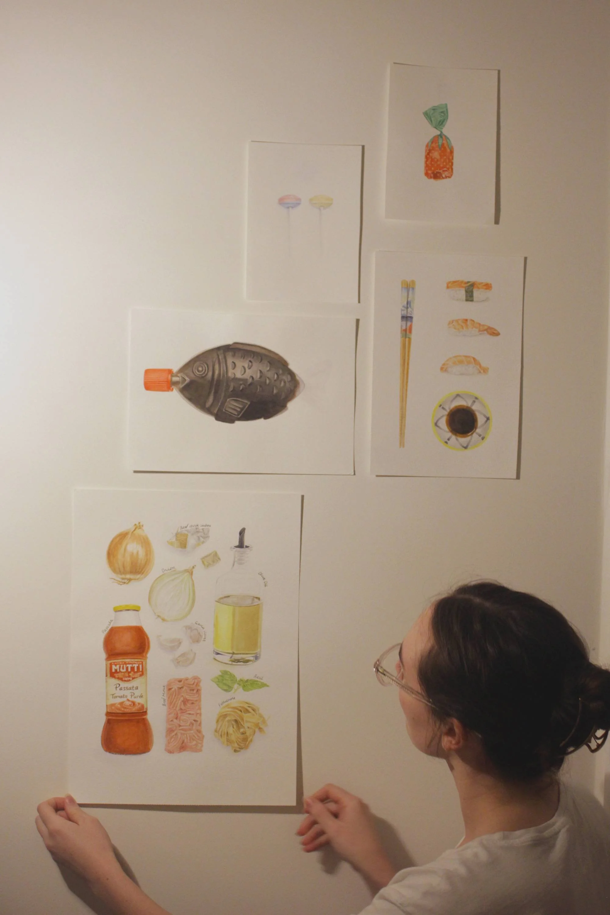 Person arranging food-themed art pieces on a wall, featuring illustrations of a fish-shaped soy sauce bottle, sushi with chopsticks, various food ingredients arranged as a unique artistic recipe.