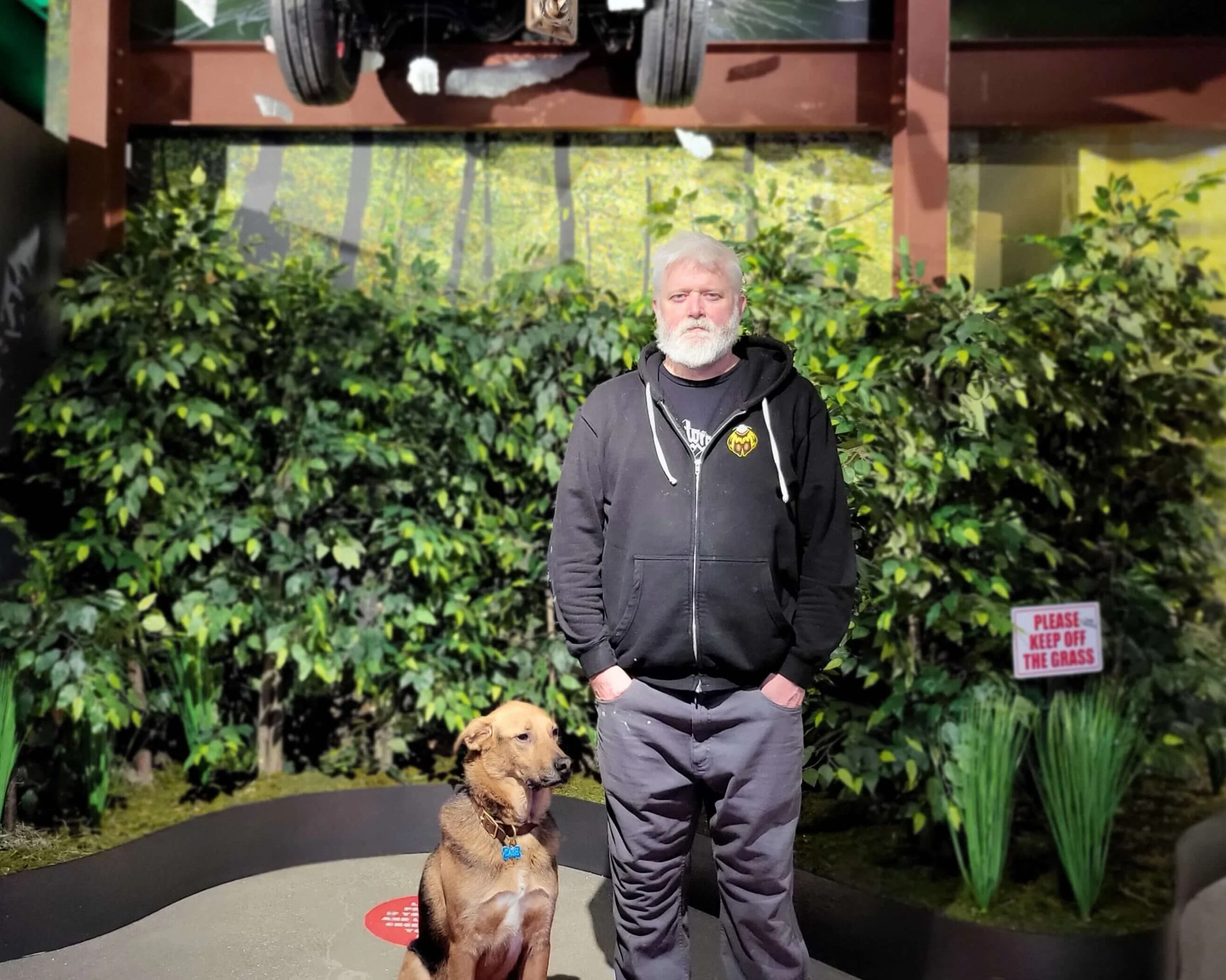 The owner Mac with white hair and beard standing with hands in pockets next to Cash, brown dog, in front of an art installation of bushes and a sign that reads "Please keep off the grass" from Ferris Bueller's Day Off.