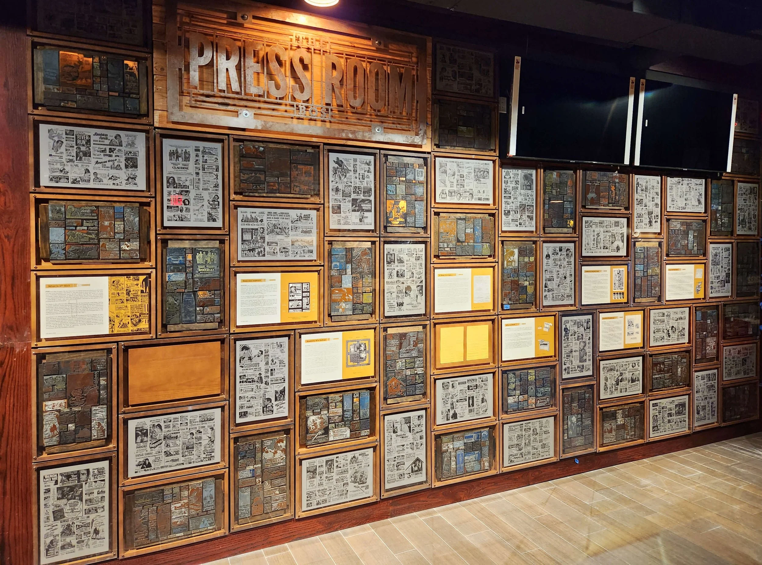 Wall with framed printing press pieces, artwork, and text displays, featuring a large wooden sign that reads 'PRESS ROOM' above the display.