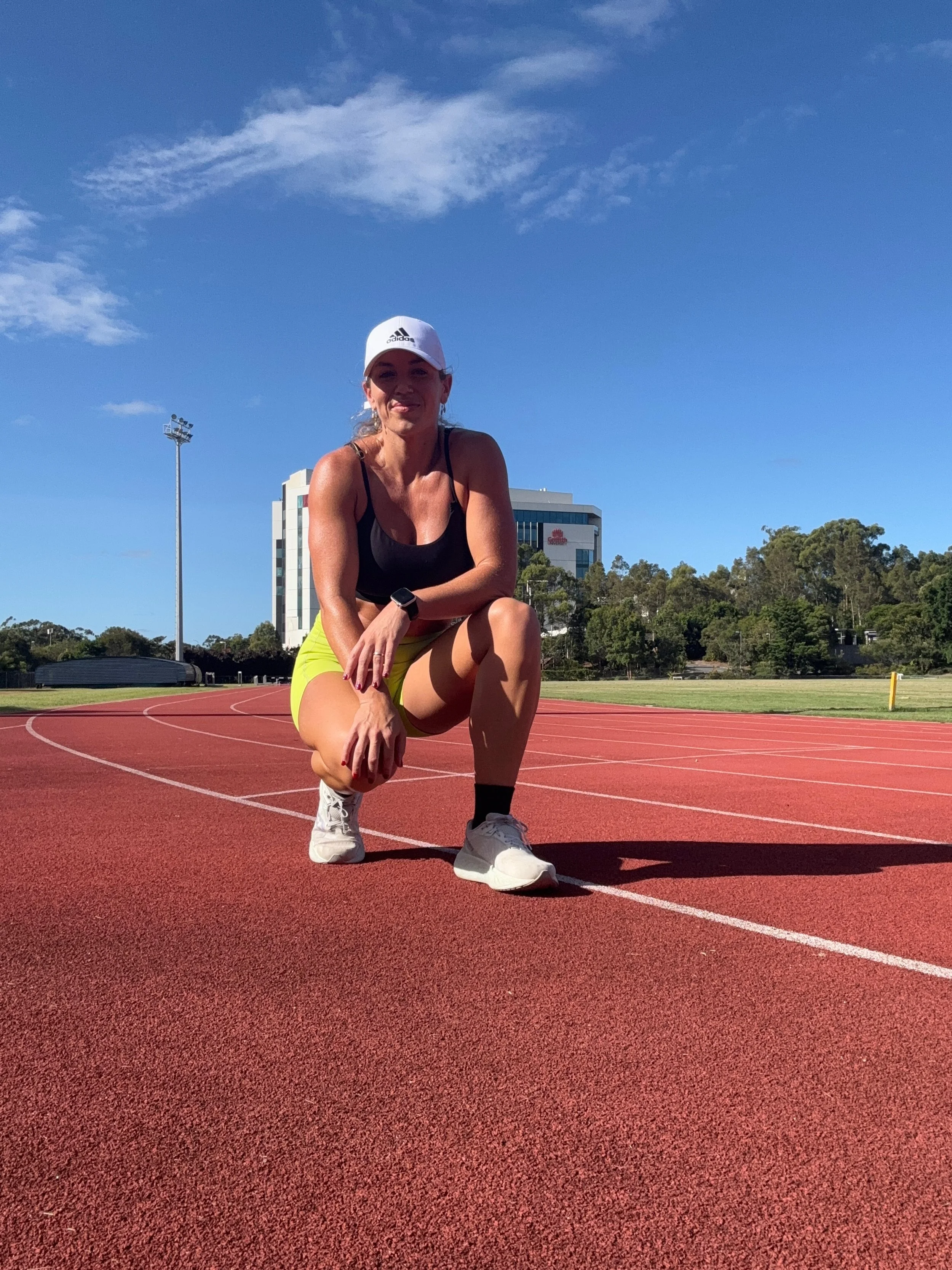 A woman in athletic clothing crouching on a red running track, smiling at the camera with a building and trees in the background on a sunny day.