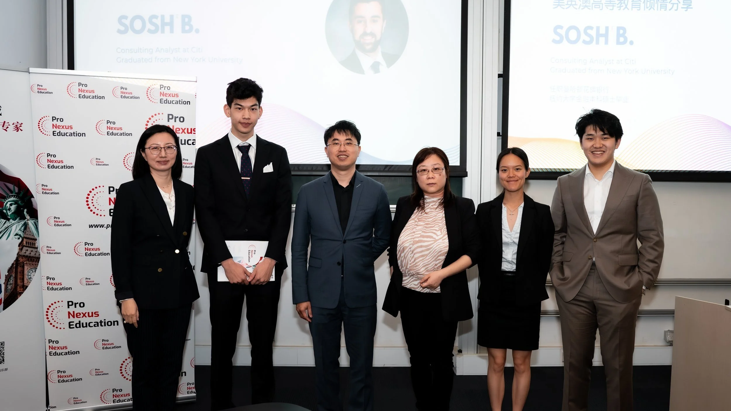 Group of six professionally dressed people standing in a row in front of presentation screens, smiling for the photo, at an educational event or conference.