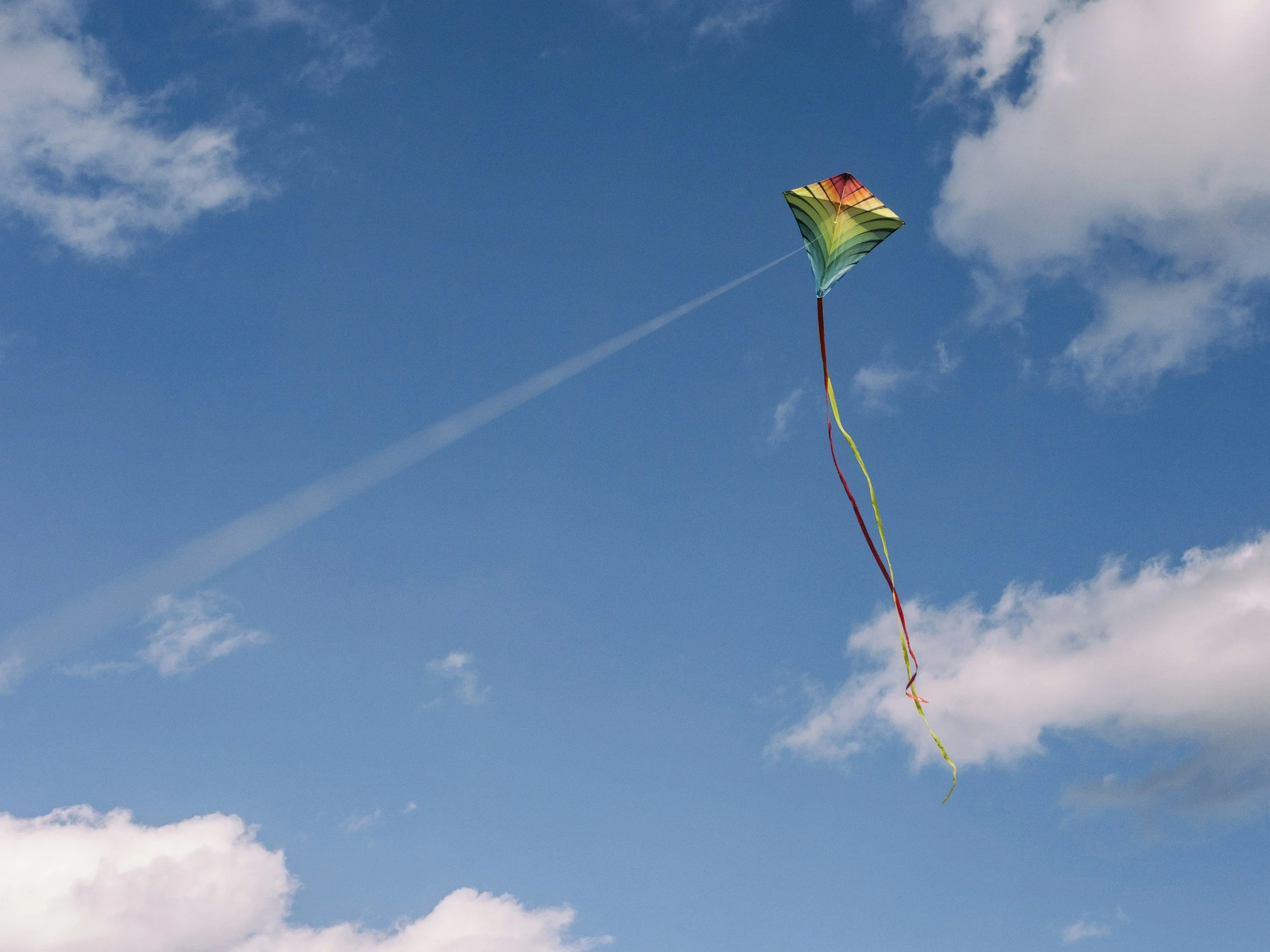 Colorful diamond-shaped kite flying in a partly cloudy blue sky, with a long tail of red and yellow ribbons.