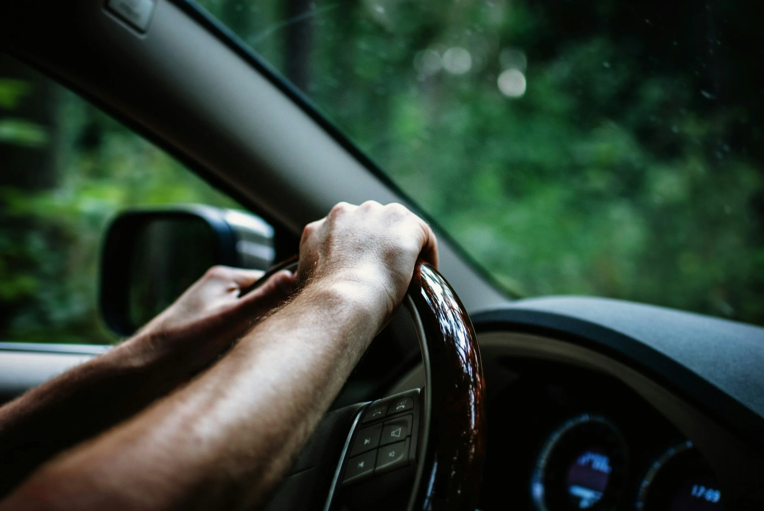 Person driving a vehicle through a wooded area, holding the steering wheel with both hands.