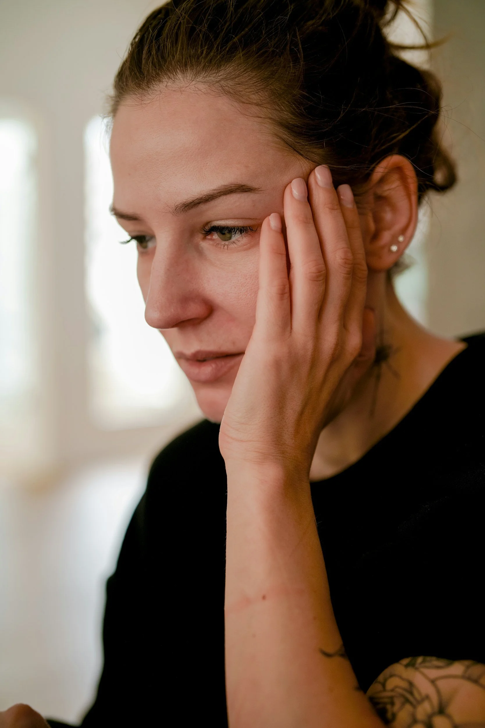 A woman with brown hair tied back, wearing earrings, with her hand on her face, looking downward with a contemplative expression.