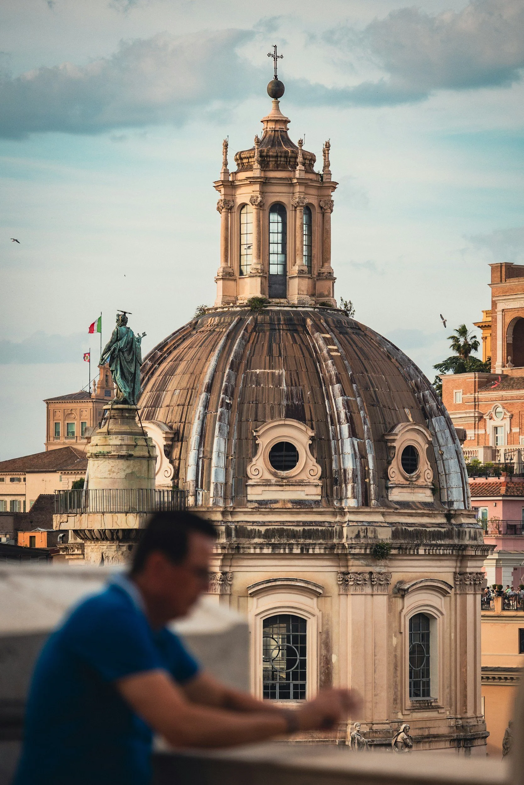 The image shows a historic building with a large, weathered dome and ornate architectural details. A man in a blue shirt and a face mask is blurred in the foreground. The sky above is partly cloudy, and there are flags and other buildings in the background.