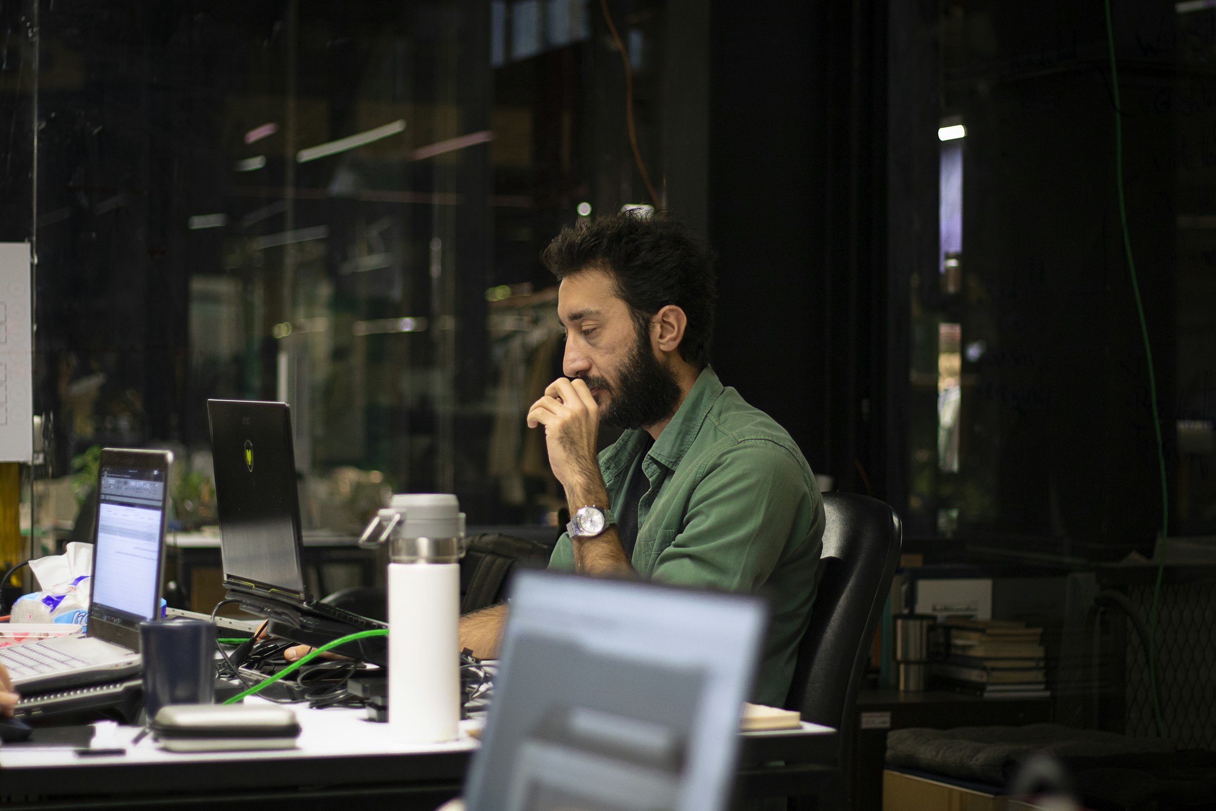 Man with a beard and dark hair, wearing a green shirt, sitting at a cluttered desk, looking at a computer screen with a thoughtful expression in a modern workspace.