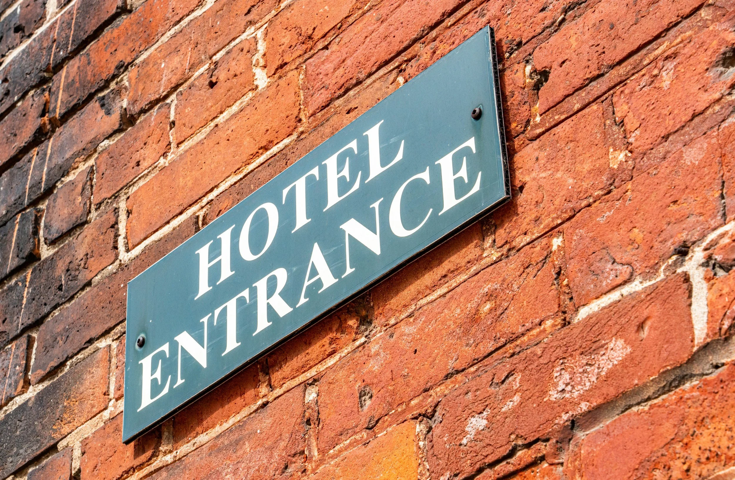 A blue street sign with white lettering that reads 'Hotel Entrance' mounted on a red brick wall.