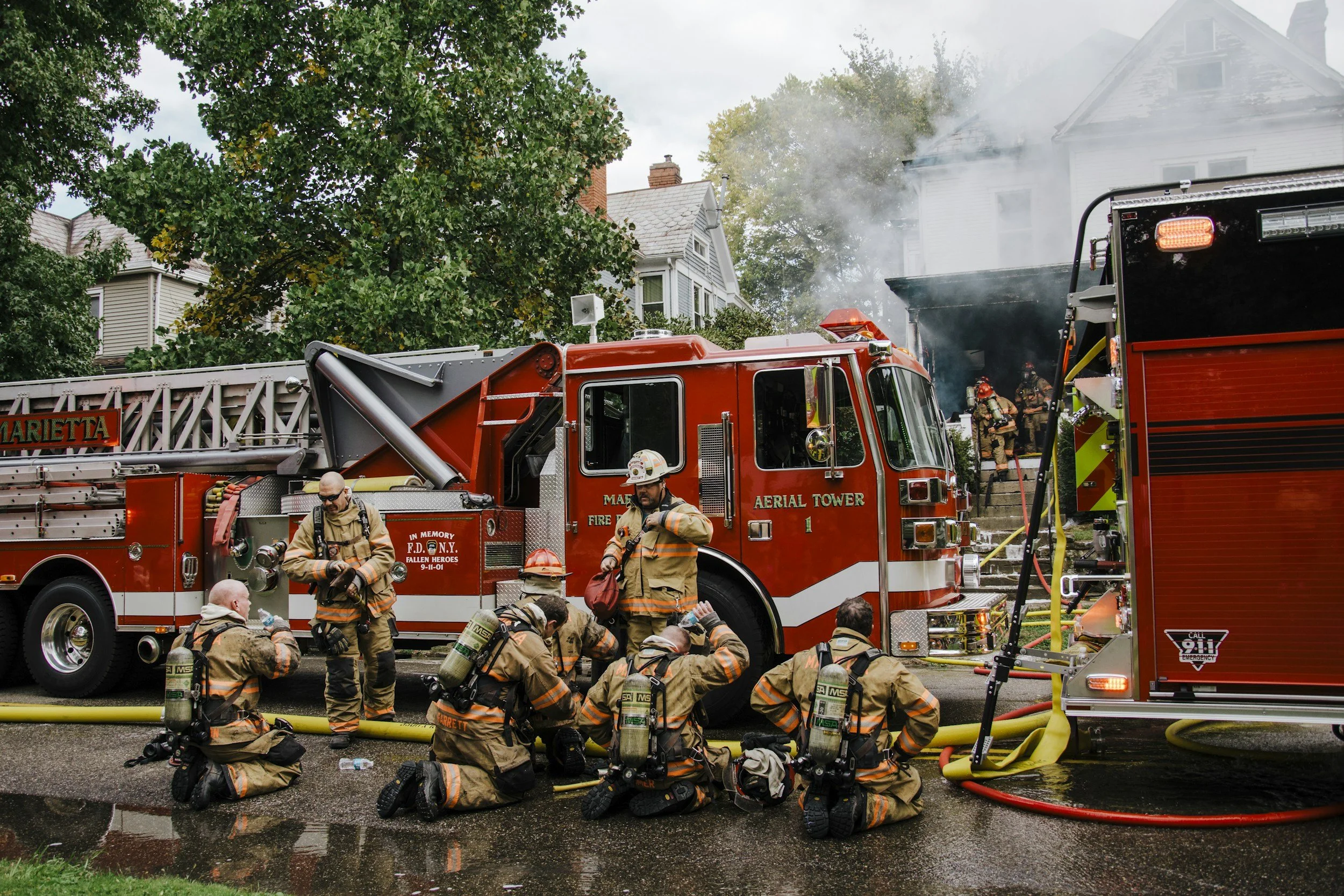 Firefighters in gear kneeling and standing in front of a red fire truck with equipment, responding to a house fire with smoke and flames visible, and firefighters working on the house steps.