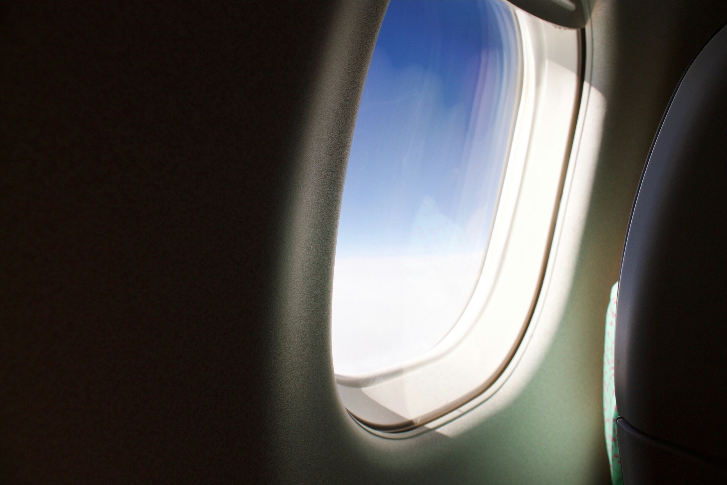 View of airplane window showing the blue sky and clouds outside, with part of the airplane's interior visible around the window.