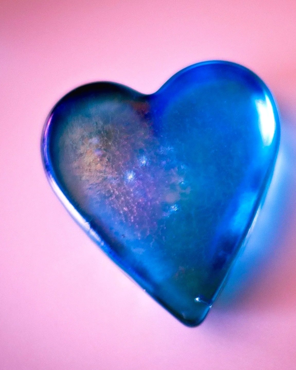 A shiny, metallic, dark blue heart-shaped object placed on a pink background.