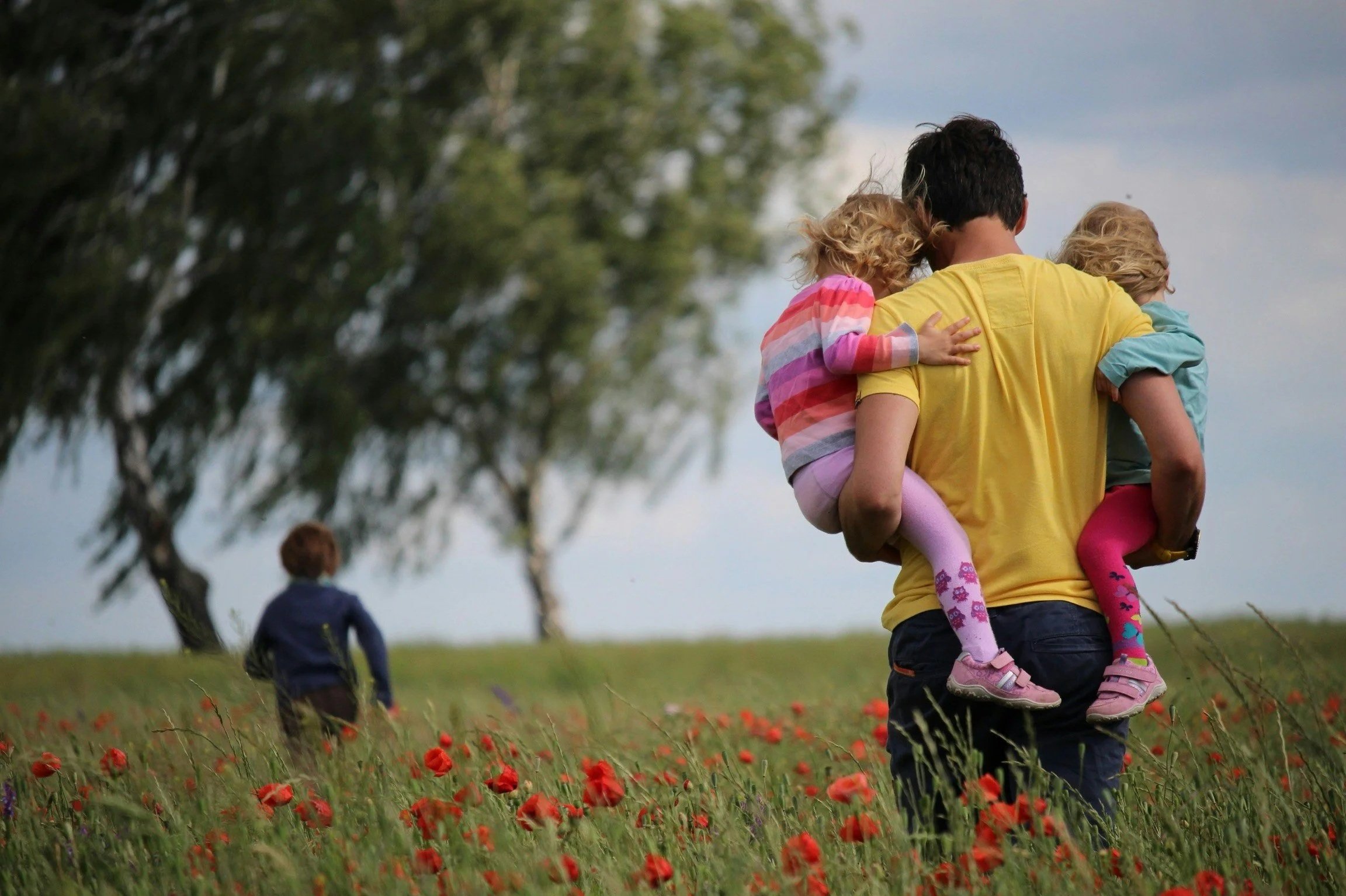A man carrying two young girls on his shoulders in a field of red poppies, with another child walking ahead in the background near trees.